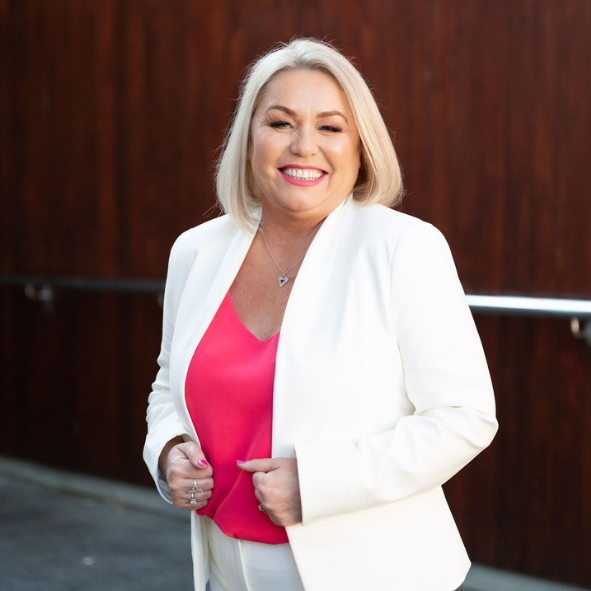 Annmarie Lord wearing a white suit with a pink shirt underneath smiling at the camera in front of a wooden wall